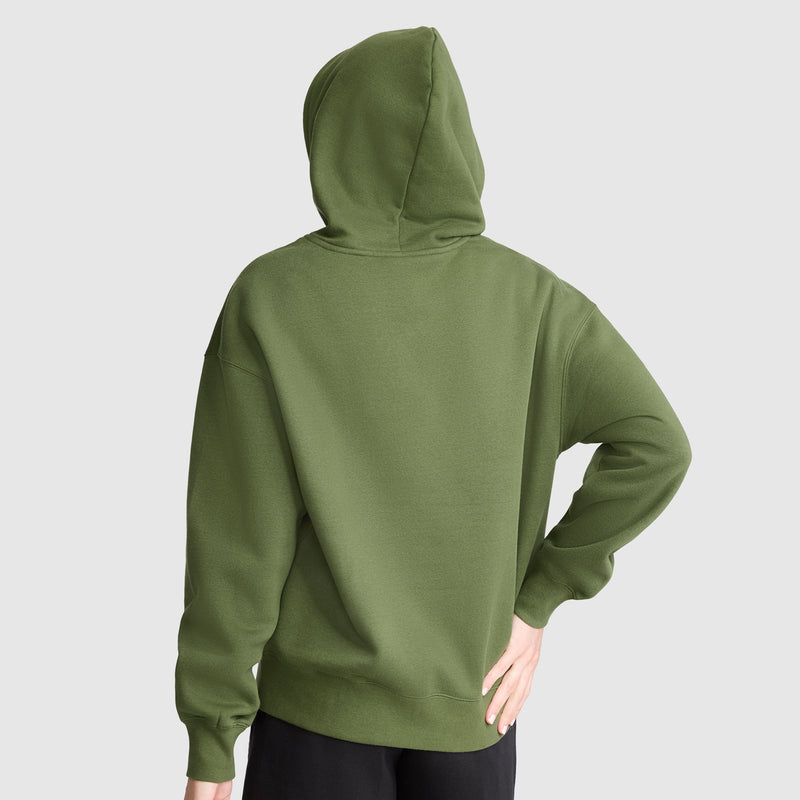 A person wearing a green Champion Tonal Script Freshman Powerblend Hoodie stands with their back to the camera, left hand by their side and right hand resting on their hip, against a plain light gray background.