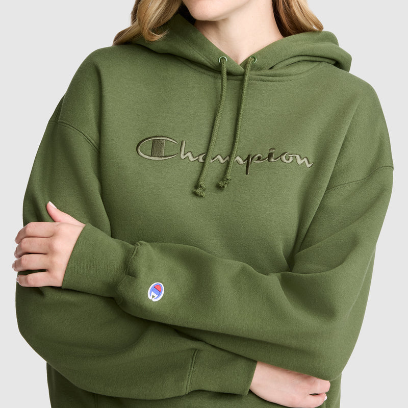 A person wears the Champion Tonal Script Freshman Powerblend Hoodie in olive green, featuring a logo on the chest. With arms folded and only the lower part of their face and torso visible, this cozy fleece sweatshirt blends comfort and style for a versatile casual look.
