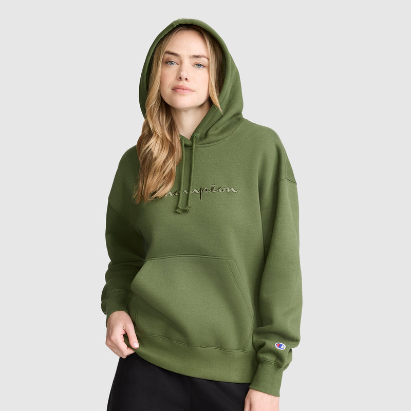 A woman wearing the Champion Tonal Script Freshman Powerblend Hoodie in green and black pants stands against a light gray background, her left hand tucked into the hoodie pocket and her right hand relaxed at her side. This cozy sweatshirt combines comfort and style.