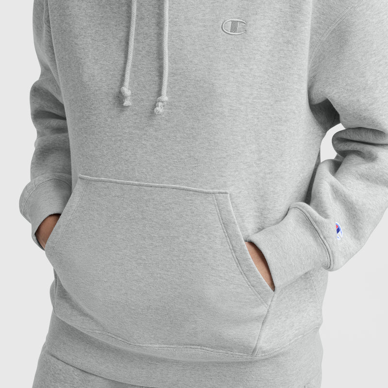 A person wears a light gray Champion Small C Freshman Powerblend Hoodie, made from midweight fleece with an adjustable drawcord hood, front pocket, and small embroidered logos on the chest and sleeve.