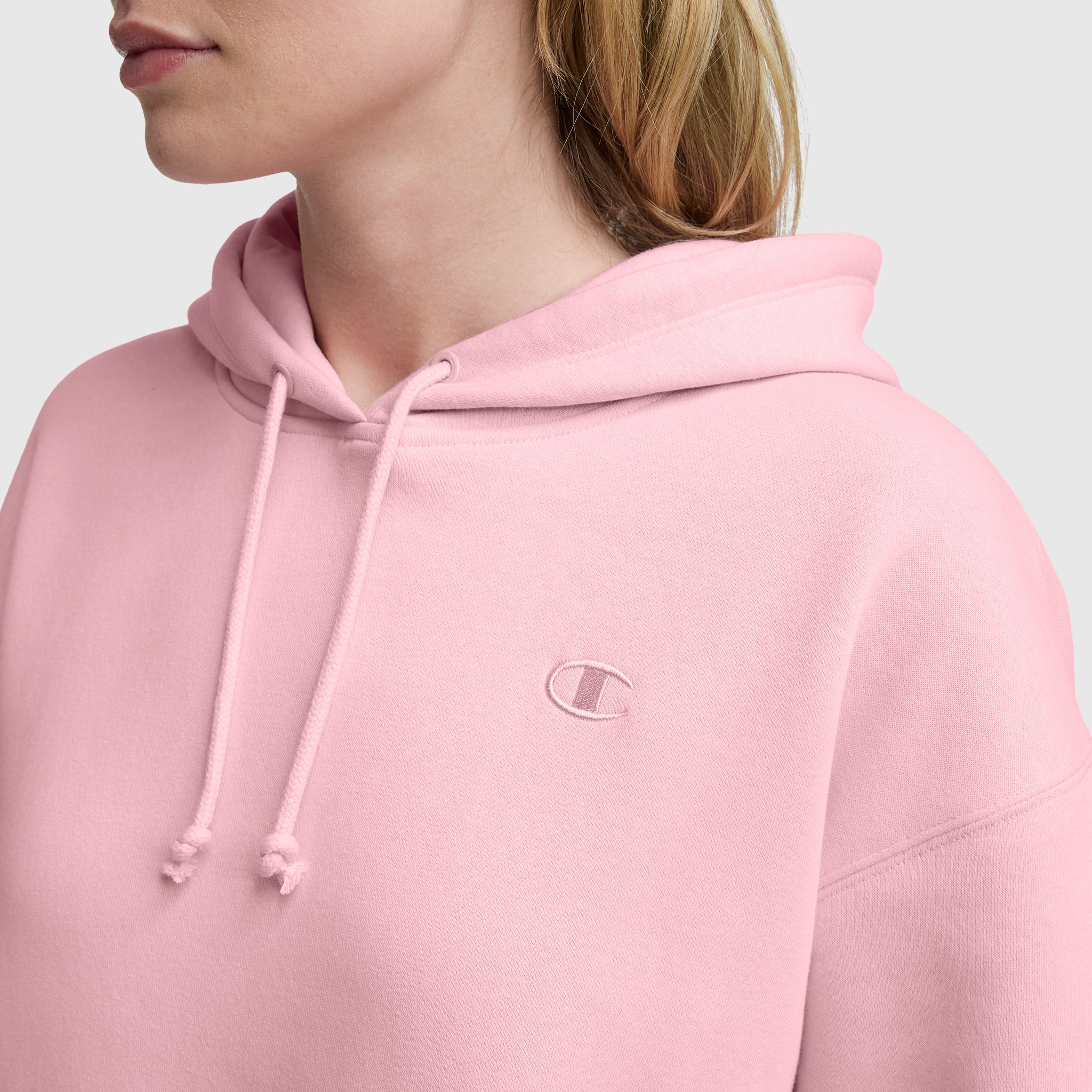 A person in a light pink Champion Small C Freshman Powerblend Hoodie, made from midweight fleece with an adjustable drawcord hood and small embroidered C logo on the chest, shown from the shoulders up against a plain light background.