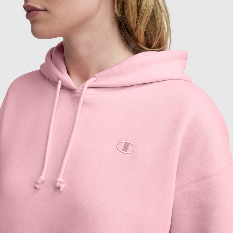 A person in a light pink Champion Small C Freshman Powerblend Hoodie, made from midweight fleece with an adjustable drawcord hood and small embroidered C logo on the chest, shown from the shoulders up against a plain light background.