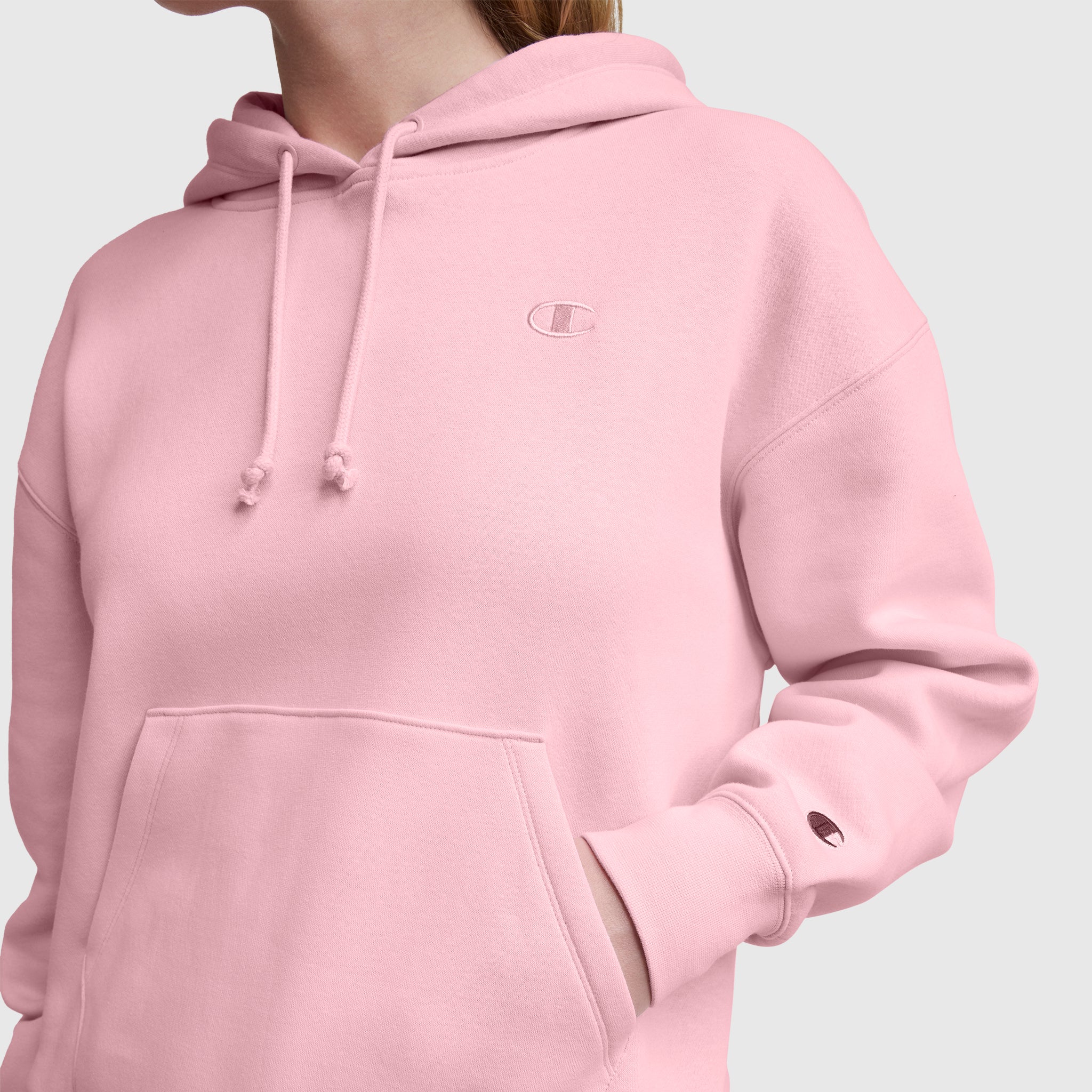 Someone wears a light pink Champion Small C Freshman Powerblend Hoodie made from midweight fleece, with their left hand in the front pocket. The adjustable drawcord hood and logos on the chest and sleeve are visible. Background: plain light gray.