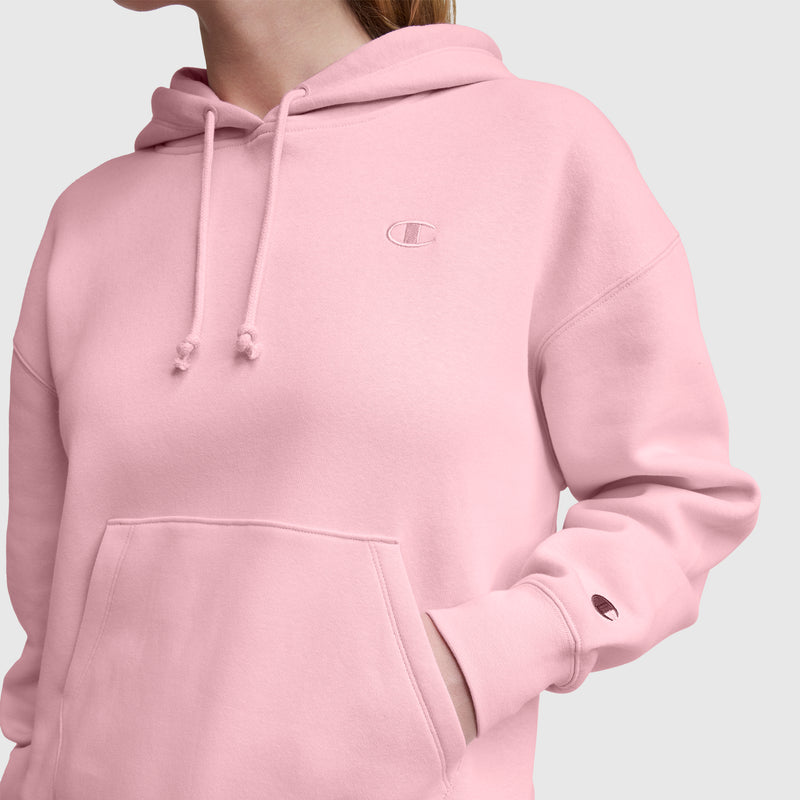 Someone wears a light pink Champion Small C Freshman Powerblend Hoodie made from midweight fleece, with their left hand in the front pocket. The adjustable drawcord hood and logos on the chest and sleeve are visible. Background: plain light gray.
