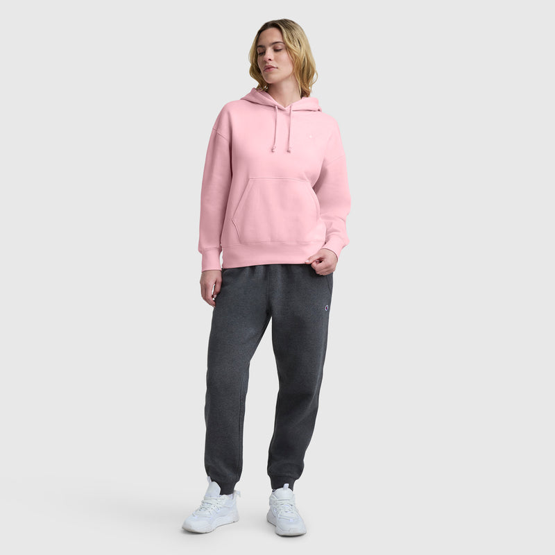 A person stands against a plain light background wearing a pink Champion Small C Freshman Powerblend Hoodie with an adjustable drawcord hood, dark gray sweatpants, and white sneakers, one hand in their pocket and looking relaxed.