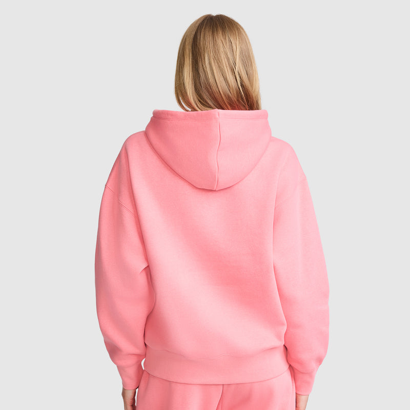 A person with long blond hair is seen from behind, wearing a bright pink Champion Small C Freshman Powerblend Hoodie and matching pants, set against a plain light grey background.