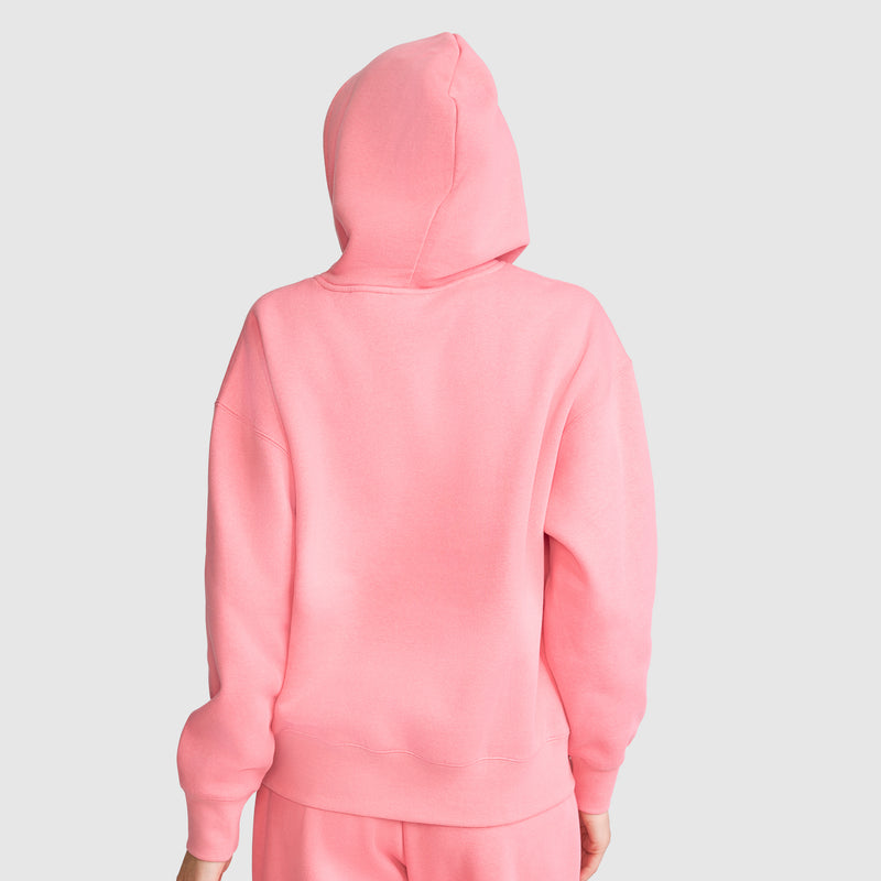 A person is wearing the Champion Small C Freshman Powerblend Hoodie in pink with the hood up, paired with matching pink pants, and facing away from the camera against a plain light gray background.