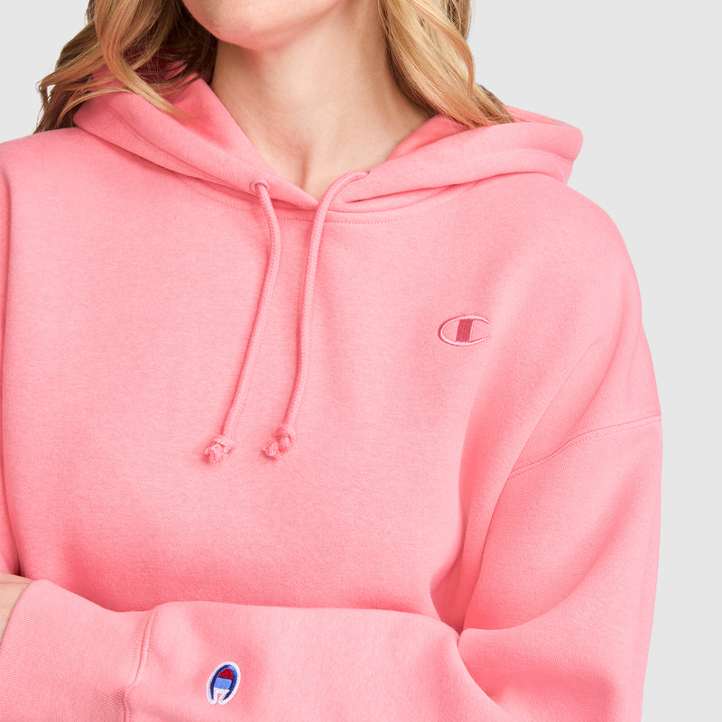 A person is shown from the shoulders down, arms crossed, wearing a pink Champion Small C Freshman Powerblend Hoodie featuring the logo on the chest and sleeve, against a plain light background.