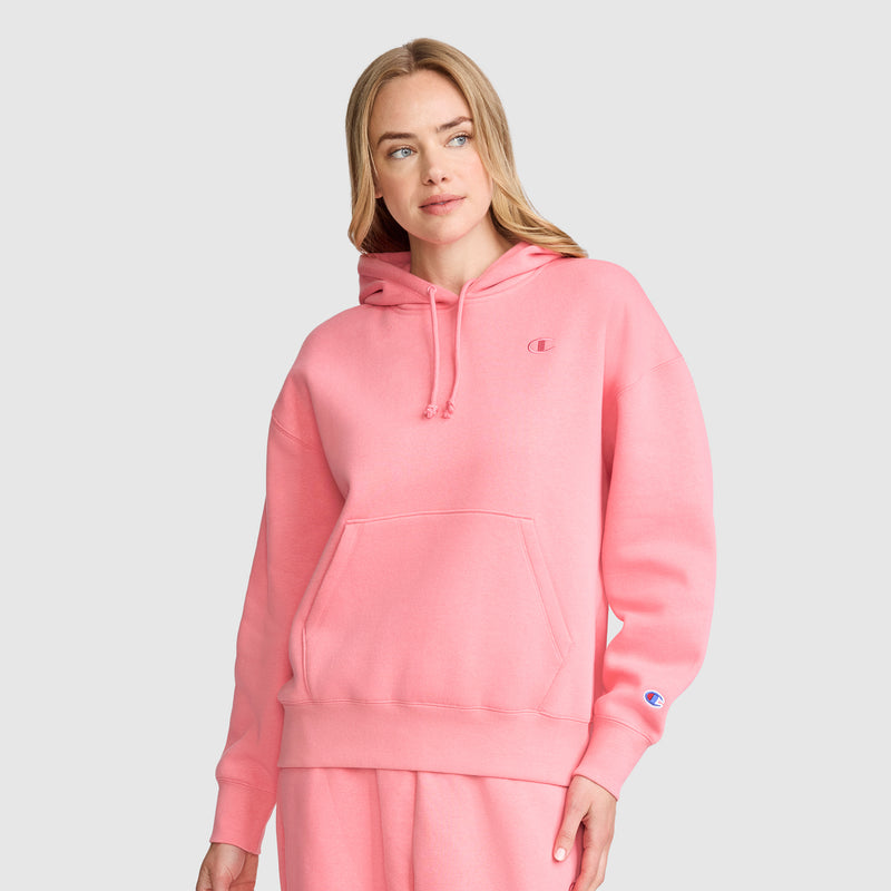 A woman is wearing the Champion Small C Freshman Powerblend Hoodie in pink, paired with matching pink sweatpants, as she stands against a plain light gray background.
