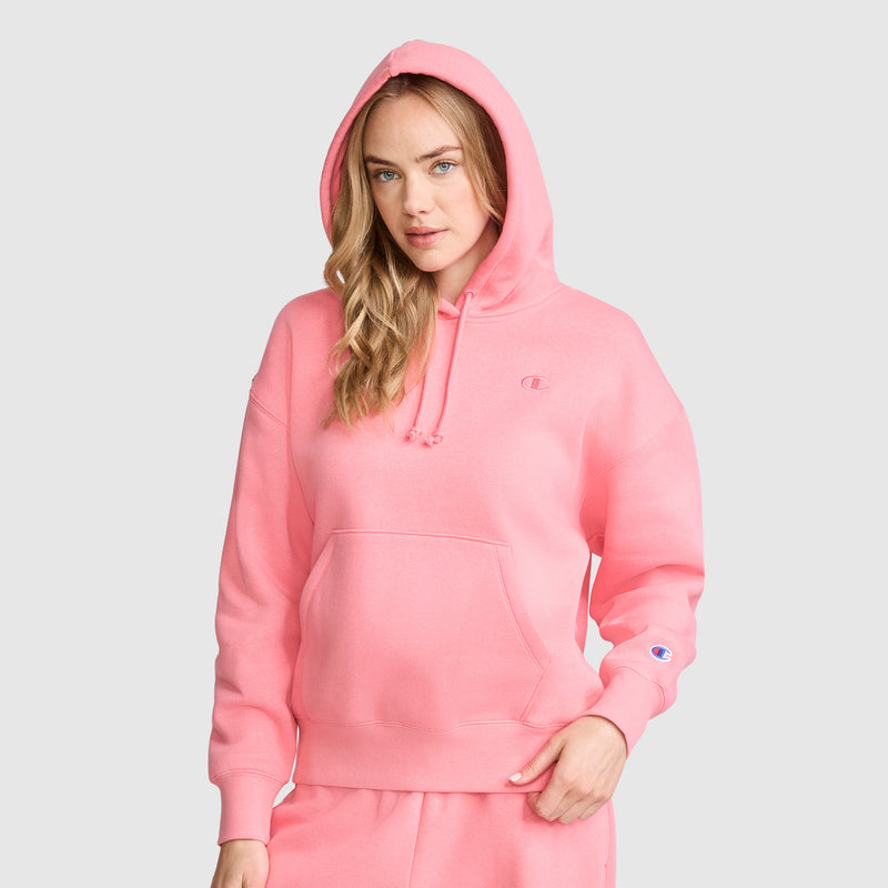 A woman wearing a bright pink Champion Small C Freshman Powerblend Hoodie and matching sweatpants stands facing forward with one hand in her pocket against a plain light gray background.