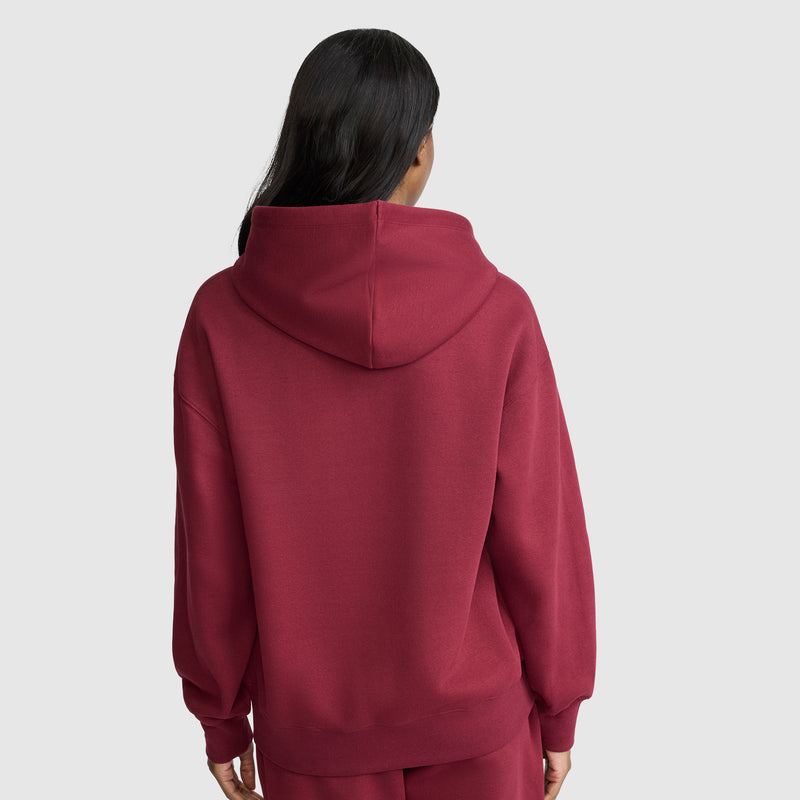 A person with long dark hair is shown from the back, wearing a loose-fitting maroon Champion Small C Freshman Powerblend Hoodie and matching pants against a plain light gray background.