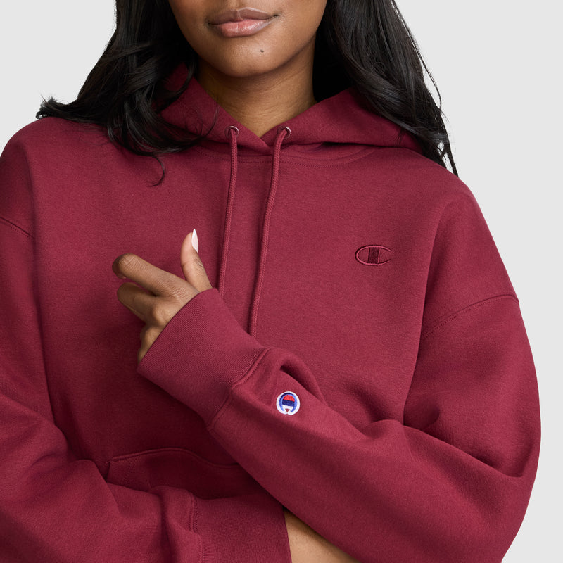 A woman wearing a maroon Champion Small C Freshman Powerblend Hoodie stands against a light background, with one hand resting on her chest and her fingernails painted white. The cozy Powerblend fleece fabric adds extra comfort to this stylish women’s hooded sweatshirt from Champion.