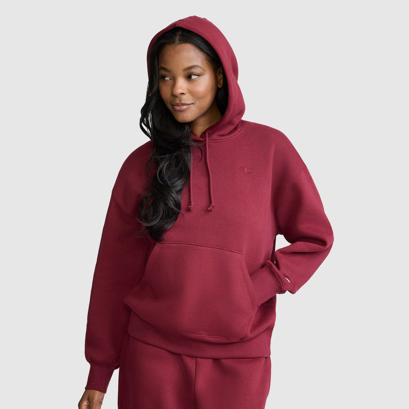 Wearing a maroon Champion Small C Freshman Powerblend Hoodie, a woman stands against a plain light gray background, looking to her left with a slight smile.