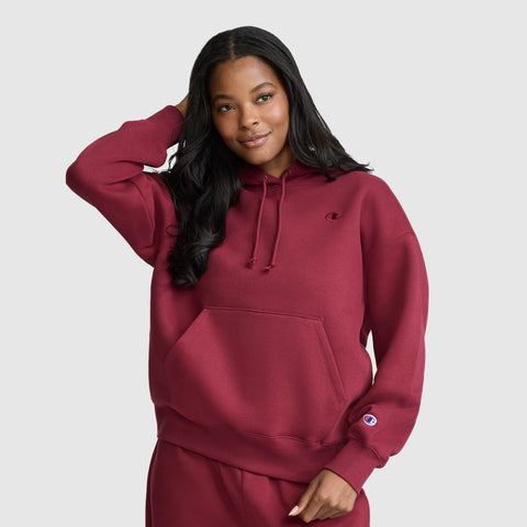 A woman wears a matching maroon Champion Small C Freshman Powerblend Hoodie and sweatpants, smiling with one hand touching her hair, standing against a plain light gray background.