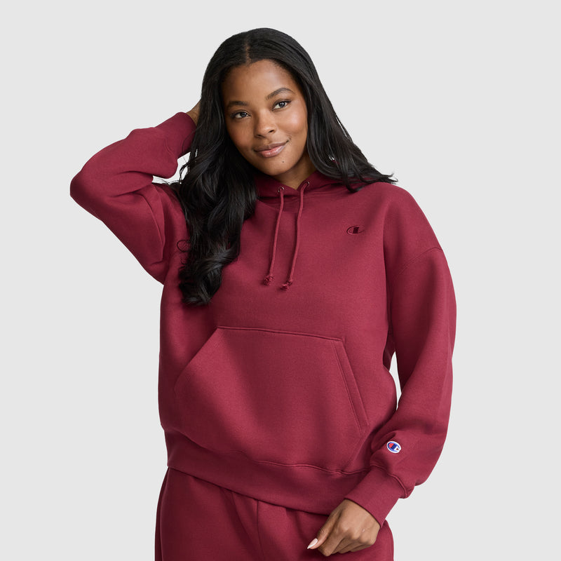 A woman wears a matching maroon Champion Small C Freshman Powerblend Hoodie and sweatpants, smiling with one hand touching her hair, standing against a plain light gray background.