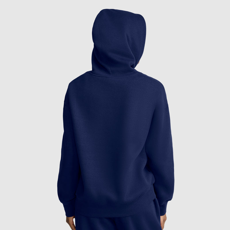 A person wearing a dark blue Champion Small C Freshman Powerblend Hoodie with the hood up, facing away from the camera against a plain light gray background. Only the back side of the hoodie and parts of the person’s hands are visible.