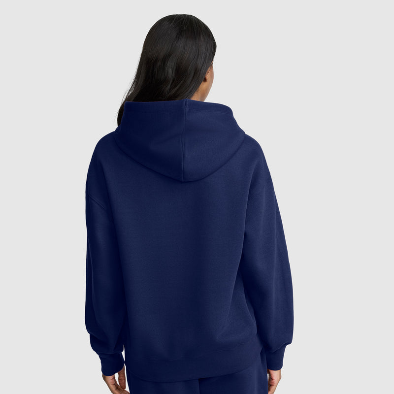 A person with long dark hair, seen from the back and wearing a navy blue Champion Small C Freshman Powerblend Hoodie, stands against a plain light gray background.