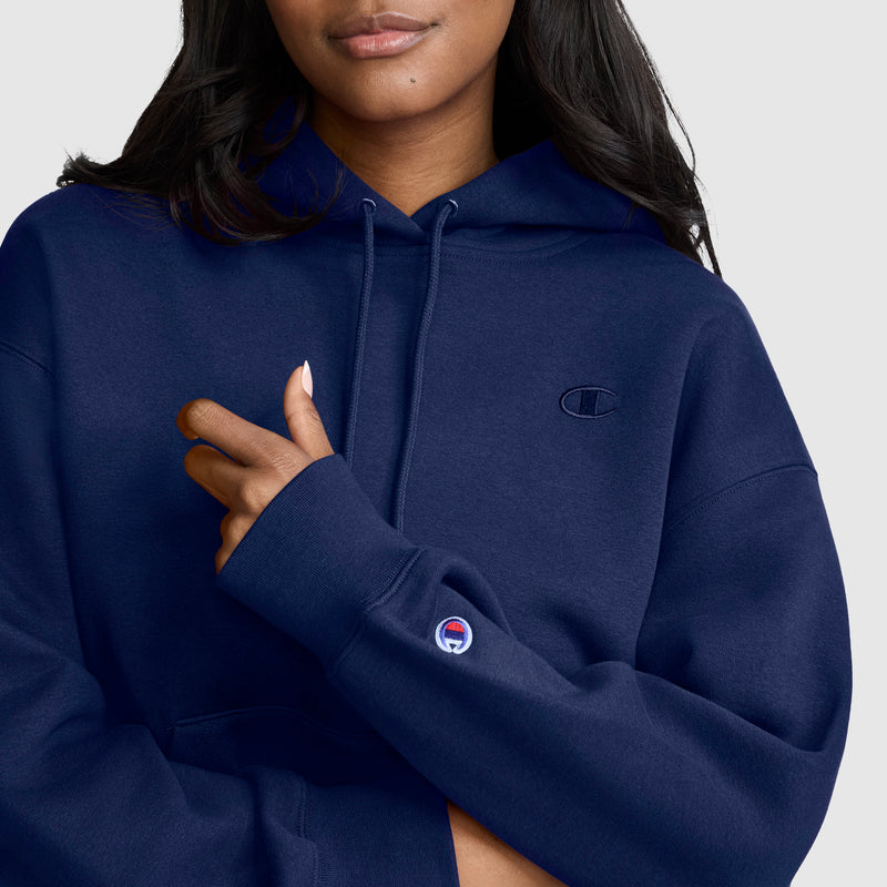 A woman wearing a navy blue Champion Small C Freshman Powerblend Hoodie with the iconic Champion logo on the chest and sleeve stands against a plain light background, one hand gently holding her sleeve.