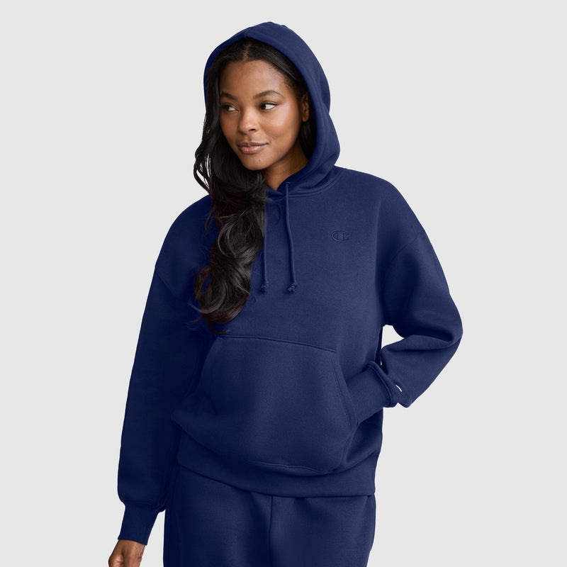 A person wearing a navy blue Champion Small C Freshman Powerblend Hoodie with a front pocket and matching pants stands against a light gray background, looking slightly to the side with a relaxed expression.