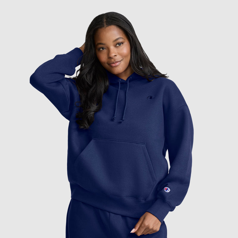 A woman with long dark hair wears a Champion Small C Freshman Powerblend Hoodie in navy blue along with matching sweatpants, standing against a light gray background and smiling as she touches her hair.
