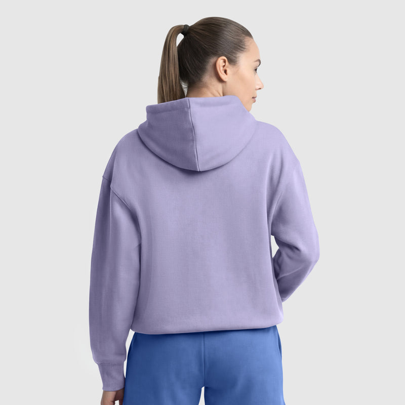 A woman with a ponytail wears the Champion Small C Freshman Powerblend Hoodie in light purple with an adjustable drawcord hood and blue pants, standing with her back to the camera against a plain light grey background.