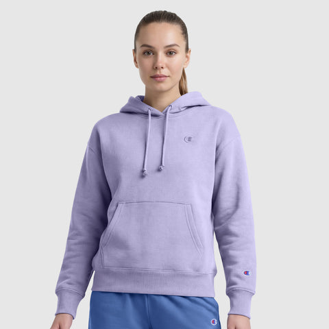 A woman in a light purple Champion Small C Freshman Powerblend Hoodie, featuring an adjustable drawcord hood and front pocket, paired with blue pants, stands facing forward against a plain light gray background.