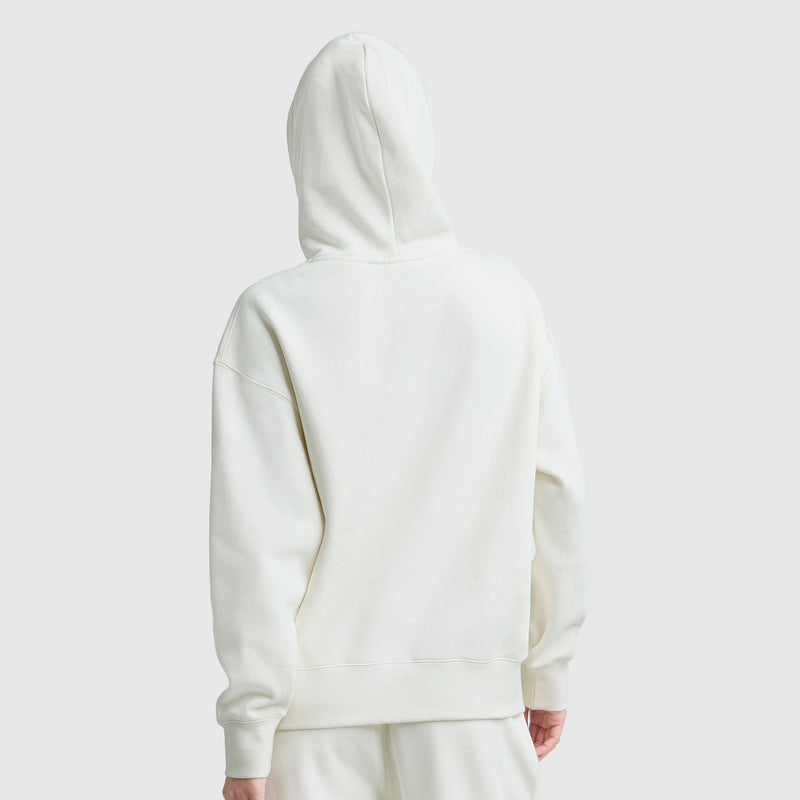 A person wearing a Champion Small C Freshman Powerblend Hoodie and matching off-white pants stands facing away from the camera against a plain light gray background, with the hood of the midweight fleece sweatshirt up, covering their head.