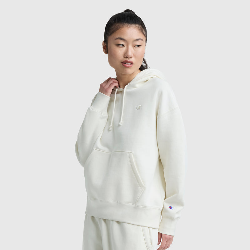 A person with long dark hair in a ponytail wears the Champion Small C Freshman Powerblend Hoodie, off-white and loose-fitting, with matching sweatpants, standing against a plain light gray background and looking slightly to the side.