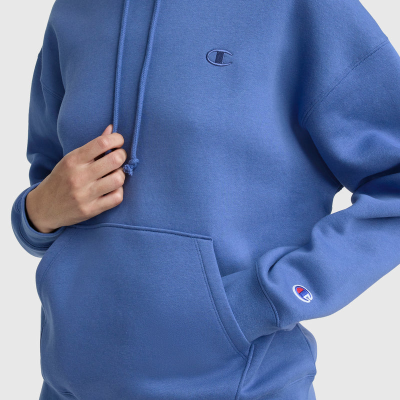 A person wears the Champion Small C Freshman Powerblend Hoodie in blue, holding the drawstring near the chest and one hand in the front pocket. The Champion logo appears on both the chest and sleeve of this midweight fleece hoodie.