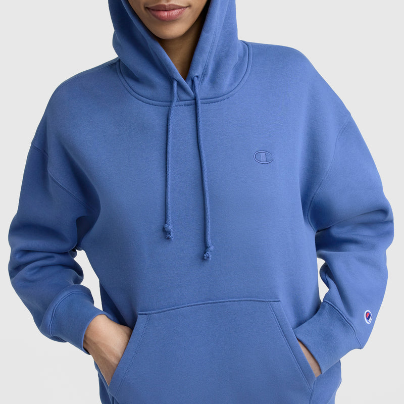 A person wears a blue Champion Small C Freshman Powerblend Hoodie with hands in the front pocket. The midweight fleece features a small embroidered logo on the chest and sleeve, set against a plain light background.