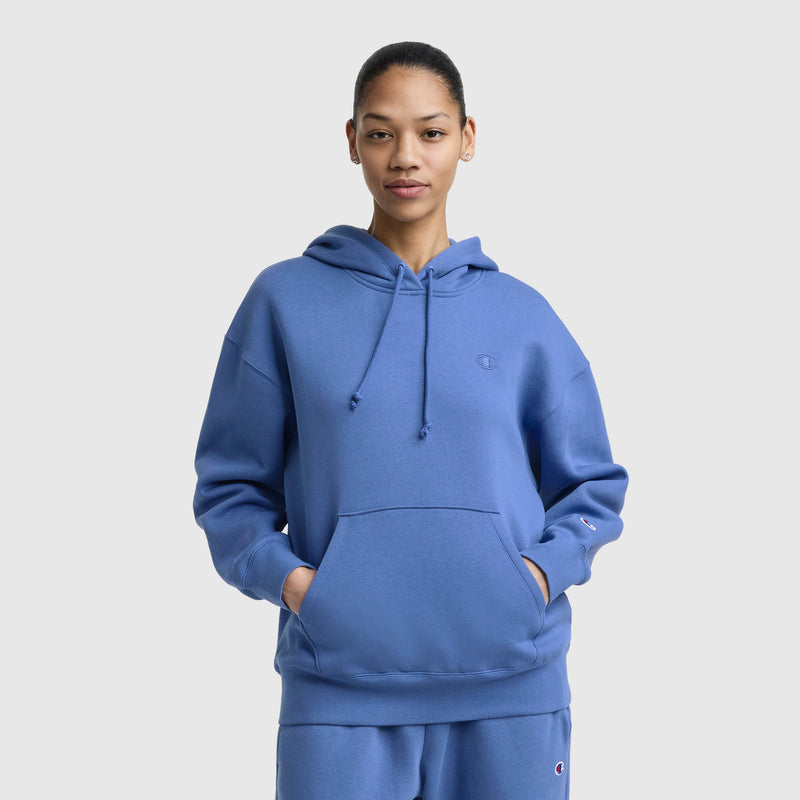 A person faces forward with hands in the front pocket, wearing a Champion Small C Freshman Powerblend Hoodie and matching blue sweatpants. They pose against a plain light gray background.