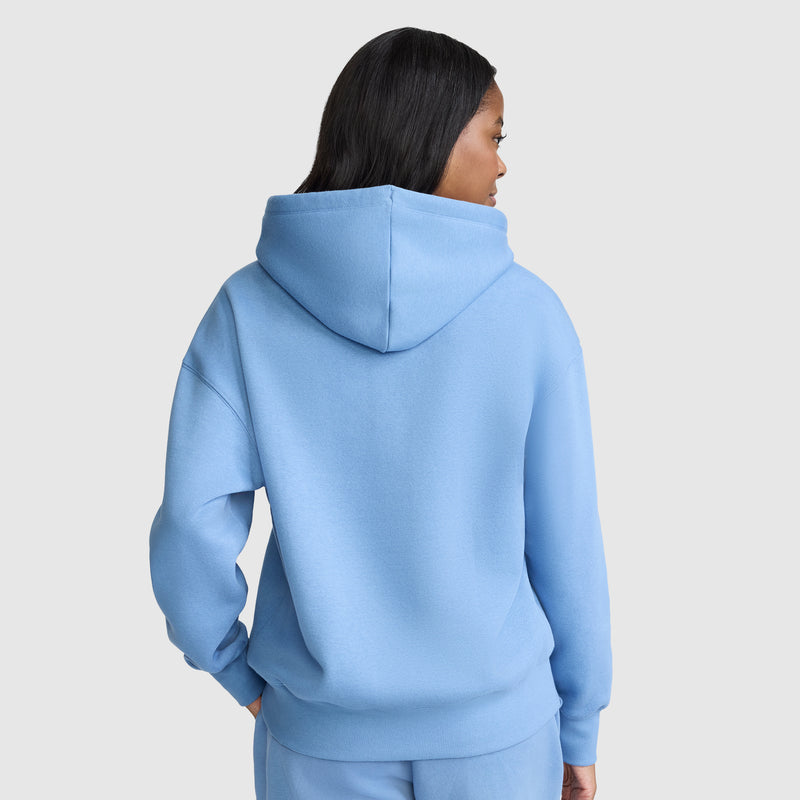 A person with long dark hair is wearing a light blue Champion Small C Freshman Powerblend Hoodie and matching pants, facing away from the camera against a plain light gray background.