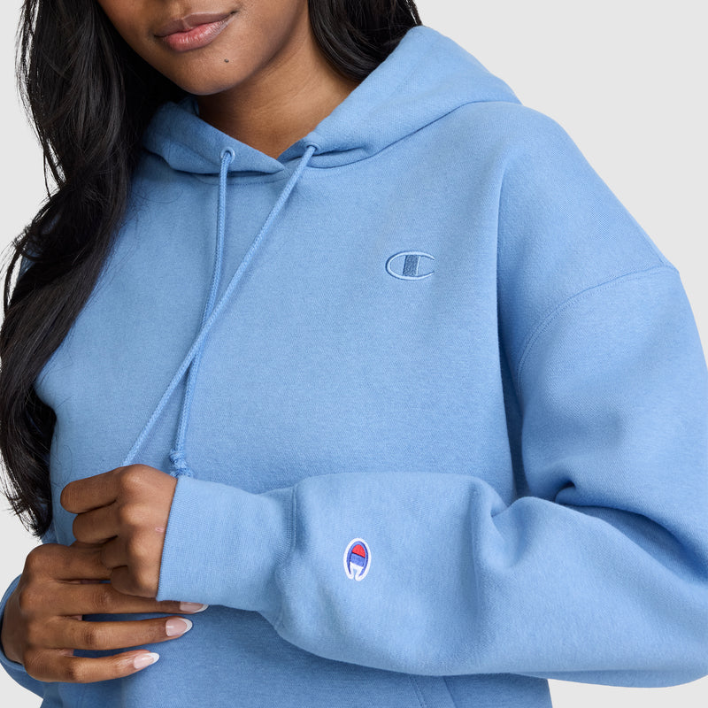 A woman wearing a light blue Champion Small C Freshman Powerblend Hoodie, featuring the Champion logo on the chest and sleeve, holds the drawstring with one hand. Only the lower half of her face is visible, emphasizing the authentic style of this classic women’s sweatshirt.