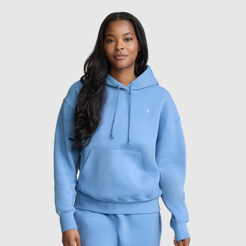 A woman with long dark hair wears a light blue Champion Small C Freshman Powerblend Hoodie and matching sweatpants, standing against a plain light gray background, embodying true Champion authenticity.