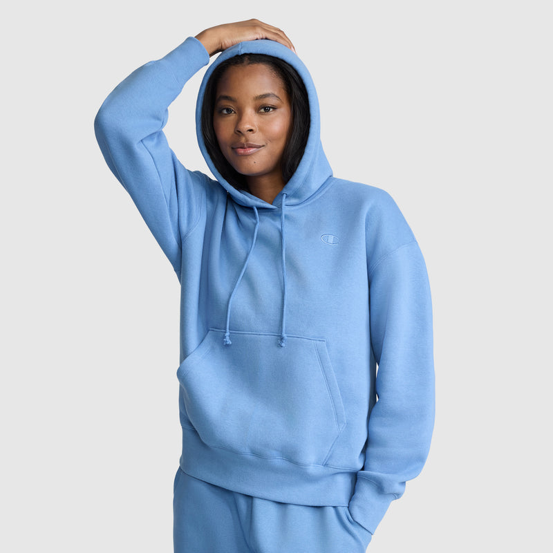 A person wearing the Champion Small C Freshman Powerblend Hoodie—a light blue women’s hooded sweatshirt paired with matching pants—stands with one hand on their head, smiling gently against a plain, light gray background, embodying true Champion style.