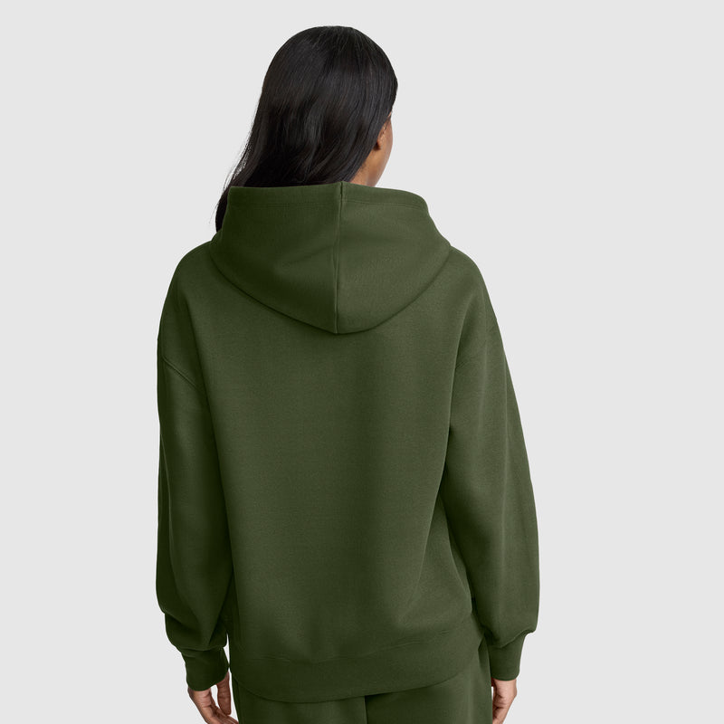 A person with long dark hair, wearing the Champion Small C Freshman Powerblend Hoodie in olive green along with matching pants, stands with their back to the camera against a plain gray background.