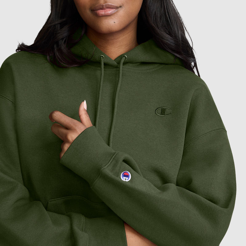 A woman wearing an olive green Champion Small C Freshman Powerblend Hoodie, with her hand holding the sleeve. This fleece hoodie features the Champion logo on both the chest and sleeve, set against a plain light-colored background.