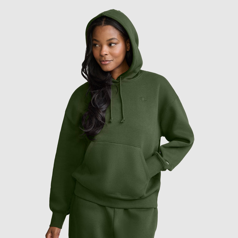 A woman wearing the Champion Small C Freshman Powerblend Hoodie in dark green, along with matching sweatpants, stands against a plain light gray background, gazing slightly to the side with a relaxed expression.
