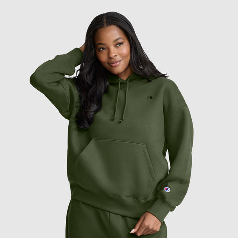 A woman wearing a Champion Small C Freshman Powerblend Hoodie and matching olive green sweatpants stands with one hand behind her head, smiling at the camera against a plain light gray background.
