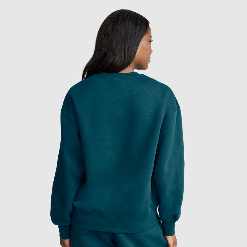 A person with long dark hair, wearing a teal Champion Powerblend Crewneck, Small “C,” and matching pants made from cozy fleece, stands facing away from the camera against a light gray background.