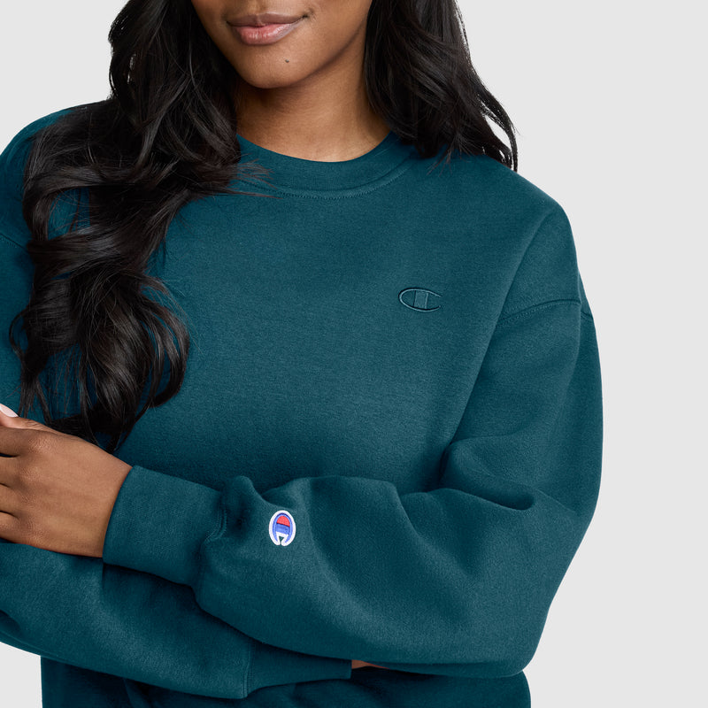 A woman with long, wavy dark hair is wearing the Champion Powerblend Crewneck, Small “C” sweatshirt in teal. This women’s crewneck features long sleeves and showcases Champion’s signature small embroidered “C” logo on the chest and sleeve. Made from soft Powerblend fleece, it delivers a comfortable and stylish fit. She is standing against a plain light background.