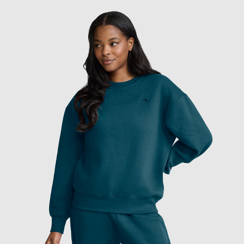 A woman with long dark hair wears a Champion Powerblend Crewneck, Small “C” sweatshirt in dark teal with matching sweatpants, standing with one hand on her hip and smiling slightly against a plain light gray background.