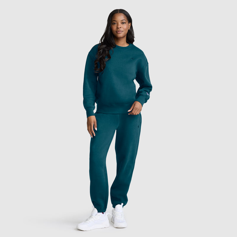 A woman stands facing forward, wearing a matching teal Champion Powerblend Crewneck, Small “C” sweatshirt and sweatpants made from cozy Powerblend fleece, paired with white sneakers, on a plain light gray background.