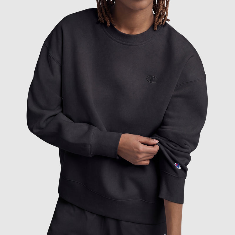 A person wears a loose-fitting black Champion Powerblend Crewneck, Small "C," and matching pants, slightly pulling up one sleeve to reveal the Champion logo patch. The background is plain light gray.