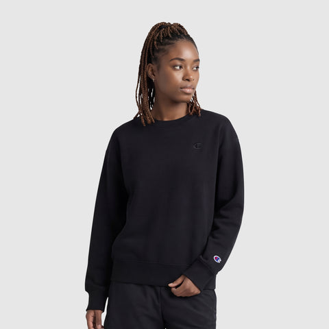 A woman with braided hair stands before a plain light gray background, wearing the Champion Powerblend Crewneck (Small "C") and black pants, looking slightly to the side with a neutral expression.