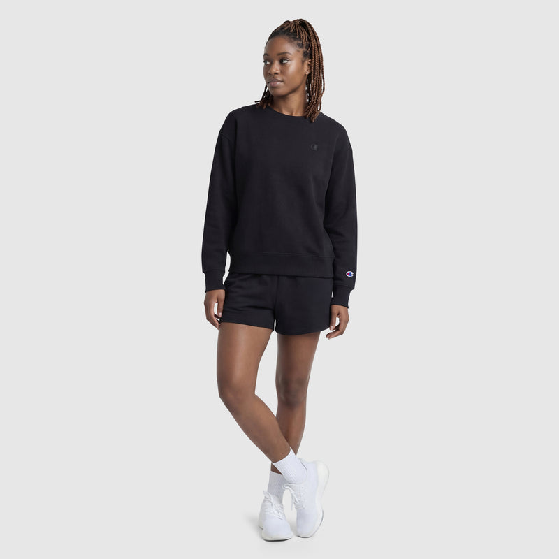 A woman in a black Champion Powerblend Crewneck with a small "C," black shorts, white socks, and white sneakers stands against a plain light gray background, looking to her left.