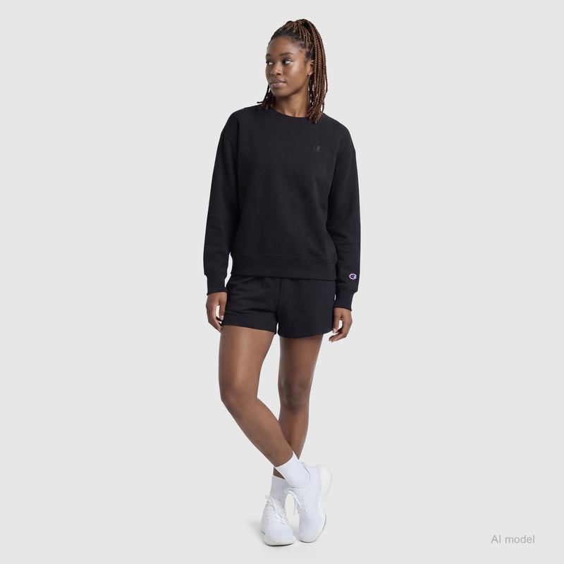 A woman stands against a plain background in a Champion Powerblend Crewneck Sweatshirt, black shorts, white socks, and sneakers. Her braided hair and relaxed posture highlight the effortless comfort of this "Small C" sweatshirt.