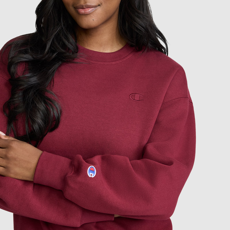 A woman wearing a maroon Champion Powerblend Crewneck, Small “C” stands against a light gray background. This women’s crewneck sweatshirt features an embroidered logo on the chest and sleeve, and is crafted from soft Powerblend fleece by Champion for ultimate comfort and style.