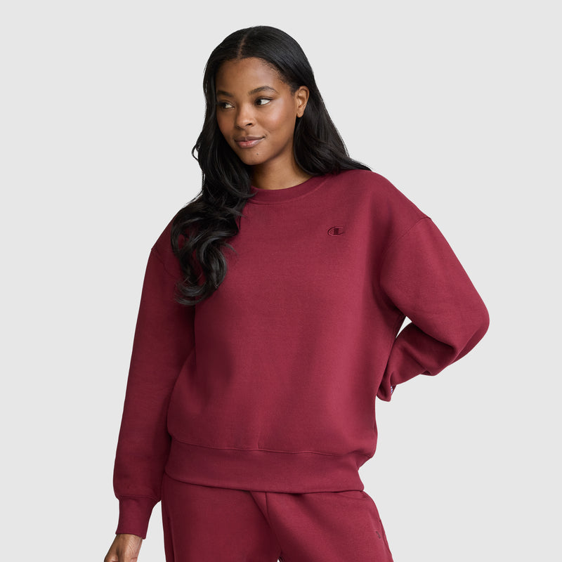 A woman with long dark hair wears a matching maroon Champion Powerblend Crewneck, Small “C”, and sweatpants, standing against a plain light gray background with one hand on her hip and a slight smile.