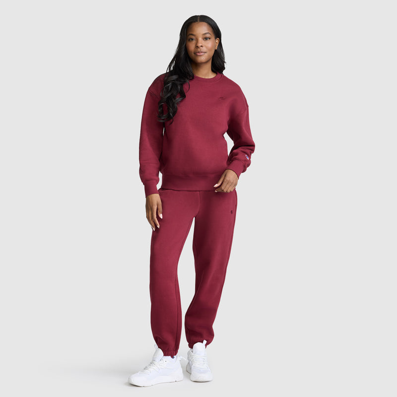 A woman stands against a plain background wearing a matching maroon Champion Powerblend Crewneck, Small “C,” paired with coordinating sweatpants and white sneakers. Her long, wavy hair and relaxed pose effortlessly highlight the comfort of the Powerblend fleece.
